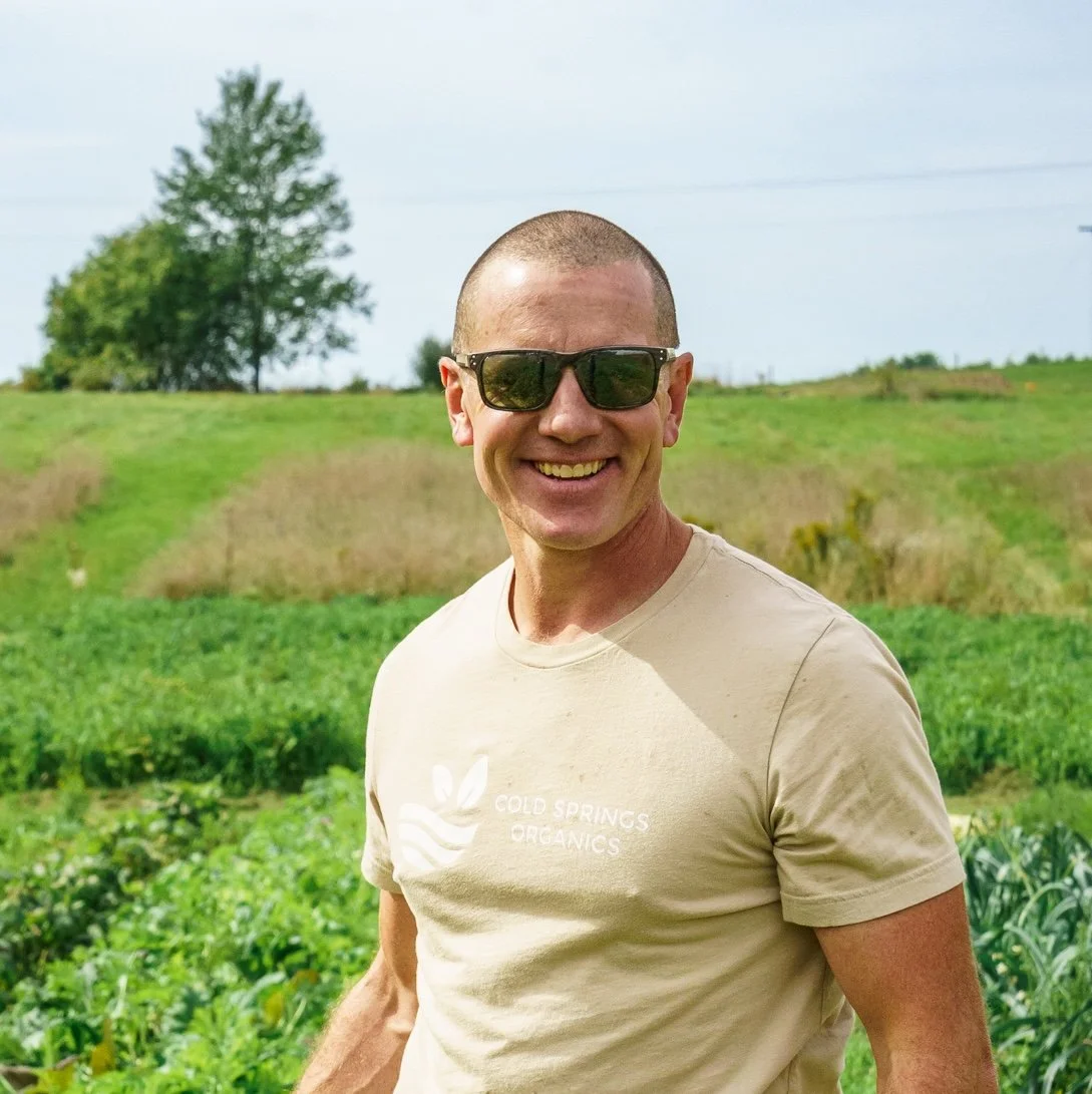 Man wearing sunglasses smiling outdoors in a green field with trees in the background.
