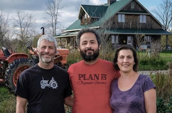 Three people standing outdoors in front of a farmhouse and a tractor, smiling at the camera.