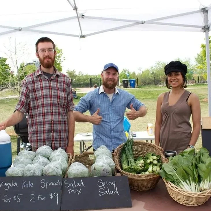 Three people standing behind a table of fresh vegetables at a farmers market, with signs showing prices for kale, spinach, and salad mix, under a white canopy outdoors.